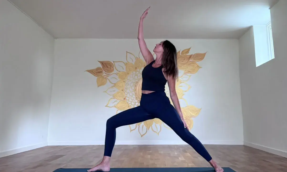 Kvinna i krigarställning i ett ljust rum med solrosmålning på väggen. En lugn yoga andrum.