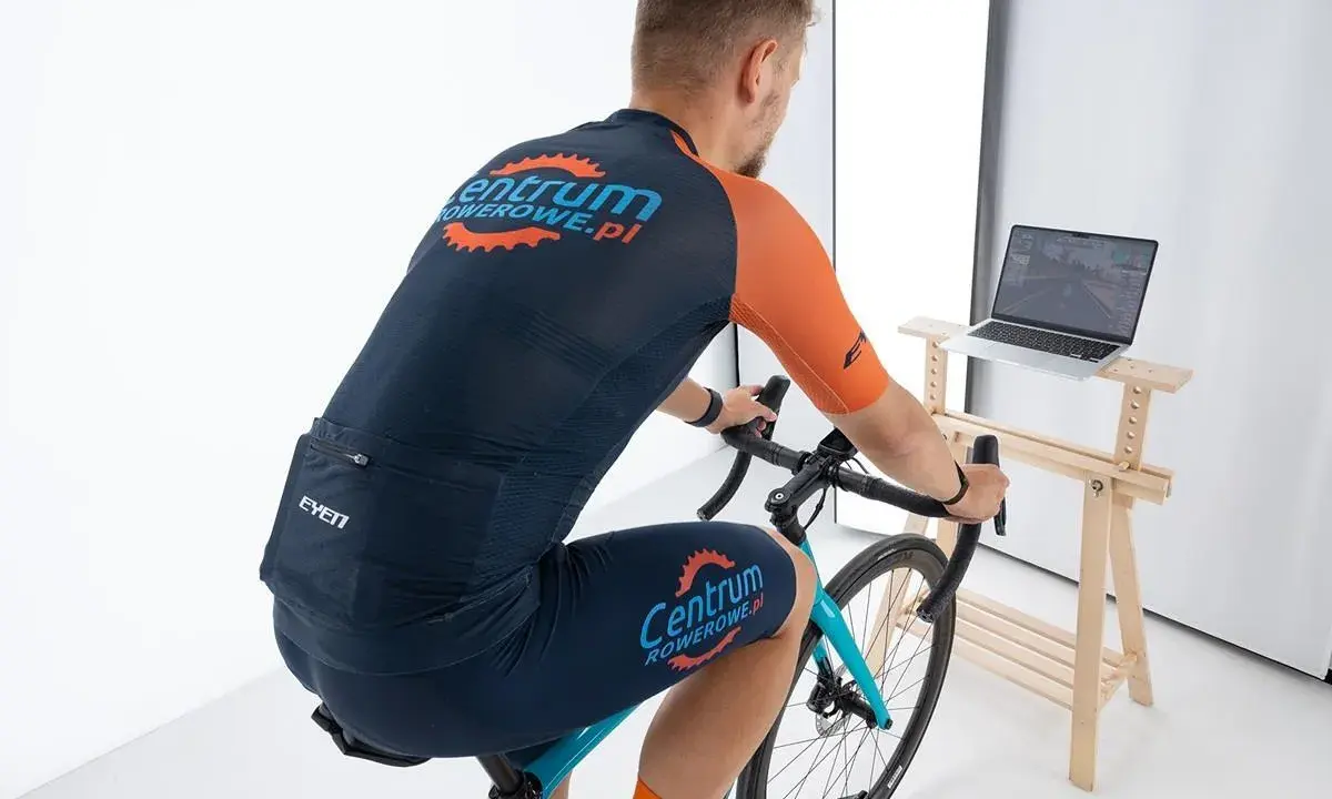 Mężczyzna w stroju kolarskim pedałuje na rowerze stacjonarnym, śledząc trasę na ekranie laptopa z aplikacją Zwift.