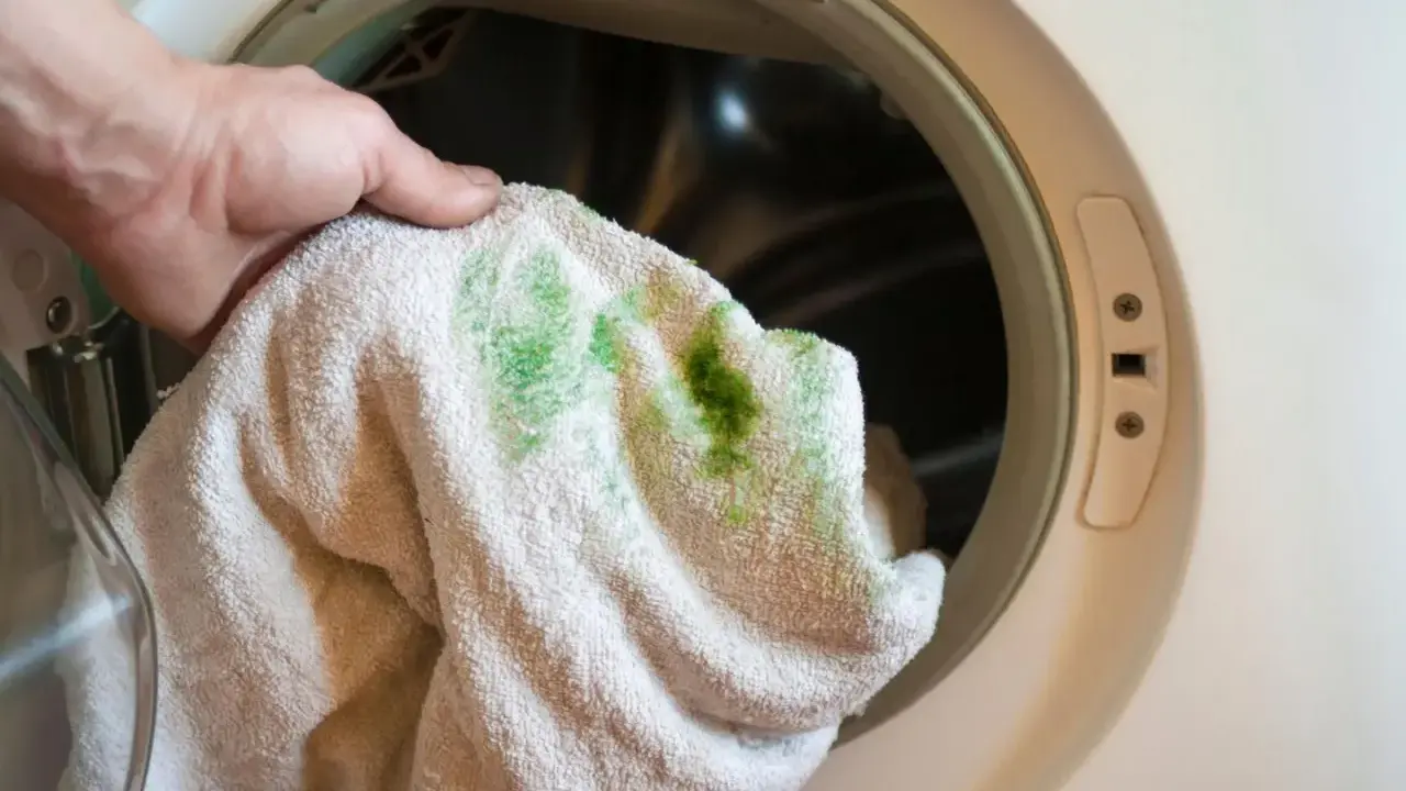 Hand hält Handtuch mit grünen, eingewaschenen Flecken vor einer Waschmaschine.