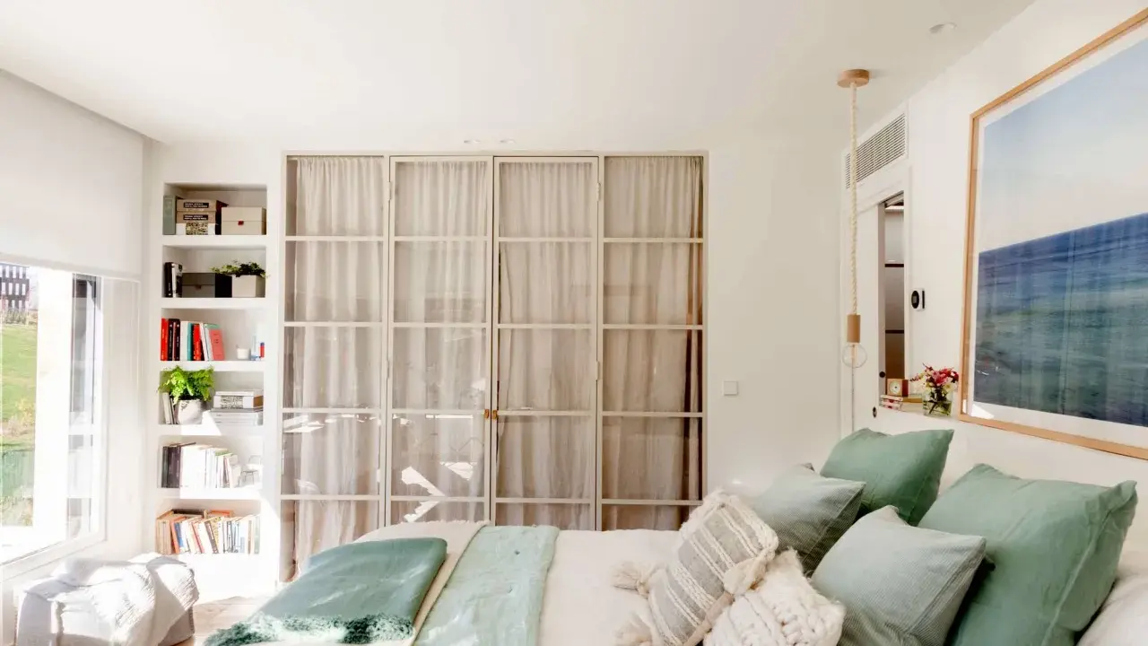 Estantería blanca aprovecha hueco entre cama y pared, con libros y plantas. Puertas de cristal con cortinas separan el espacio.