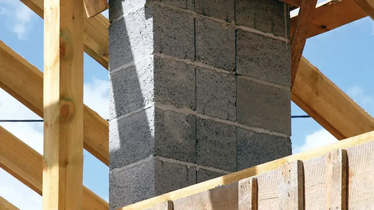 Budowa komina systemowego z bloczków betonowych, gotowego do podłączenia pieca. Drewniana konstrukcja dachu w tle.