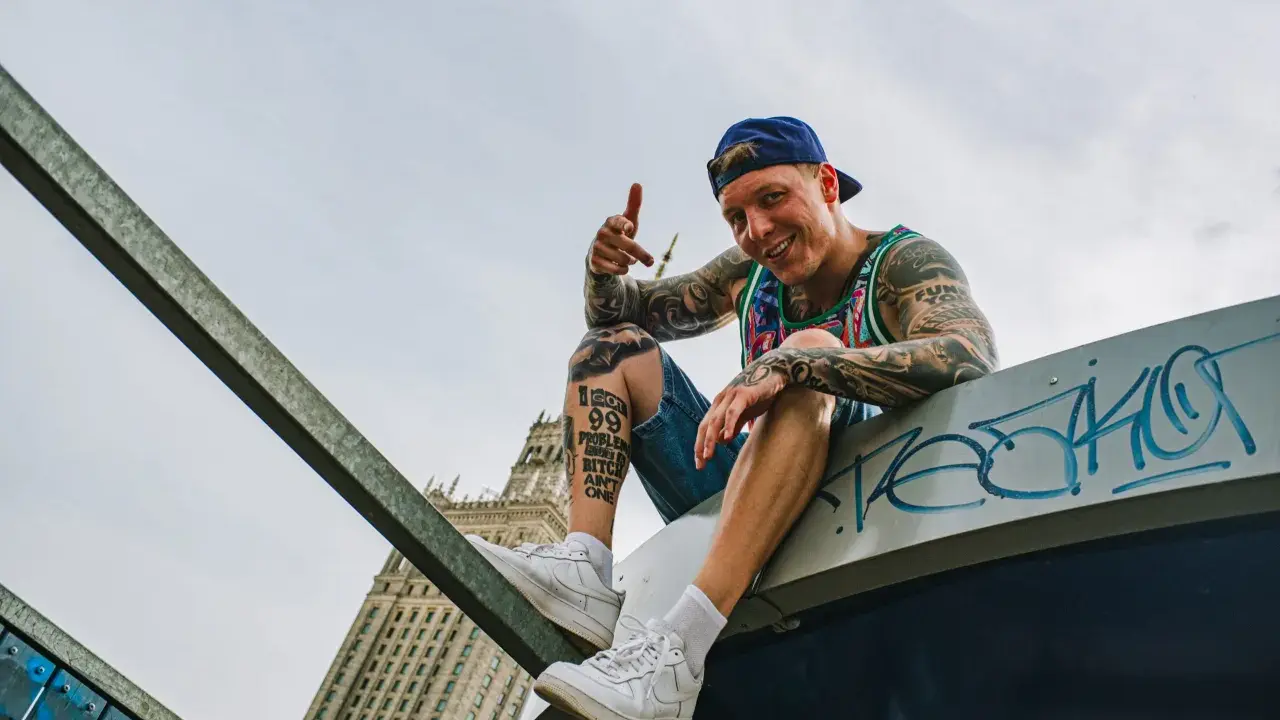 Uśmiechnięty raper z tatuażami, w czapce i kolorowej koszulce, siedzi na dachu, promując swój mixtape "sos ciuchy i borciuchy".
