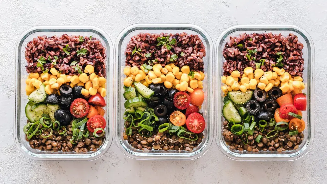 Drei Glasbehälter mit einem gesunden Essensplan für Fitness: Linsen, Gurken, Oliven, Tomaten, Mais und brauner Reis.