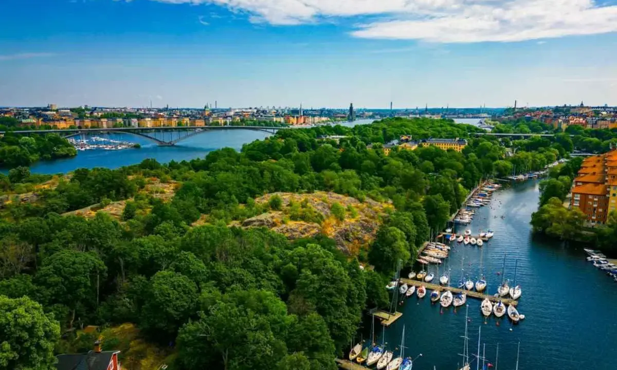 En vacker promenad i Stockholm med grönskande träd, en bro över vattnet och många segelbåtar i hamnen.
