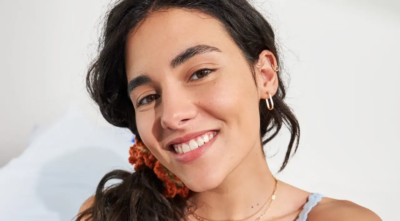 Primo piano di una giovane donna con un sorriso radioso, la sua fronte lucida riflette la luce. Indossa orecchini a cerchio dorati e una collana sottile.