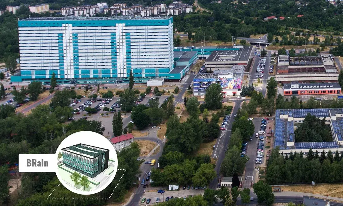 Widok z lotu ptaka na kompleks budynków, w tym duży, jasny budynek z niebieskimi akcentami, przypominający umed w Łodzi. Widać parkingi, drogi i zieleń.
