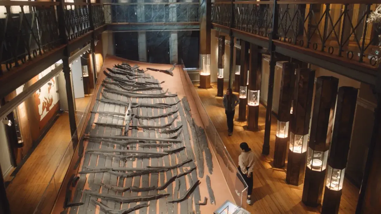 Vikingaskeppsvrak på ett museum i Göteborg. Besökare studerar utställningen med gamla trädelar.