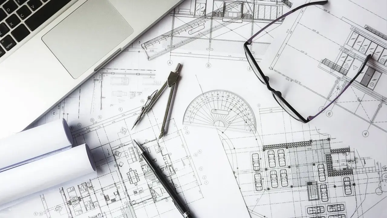 Na stole architekta leżą plany budynku, laptop, okulary i cyrkiel. To miejsce pracy nad projektem i jego kosztorysem.