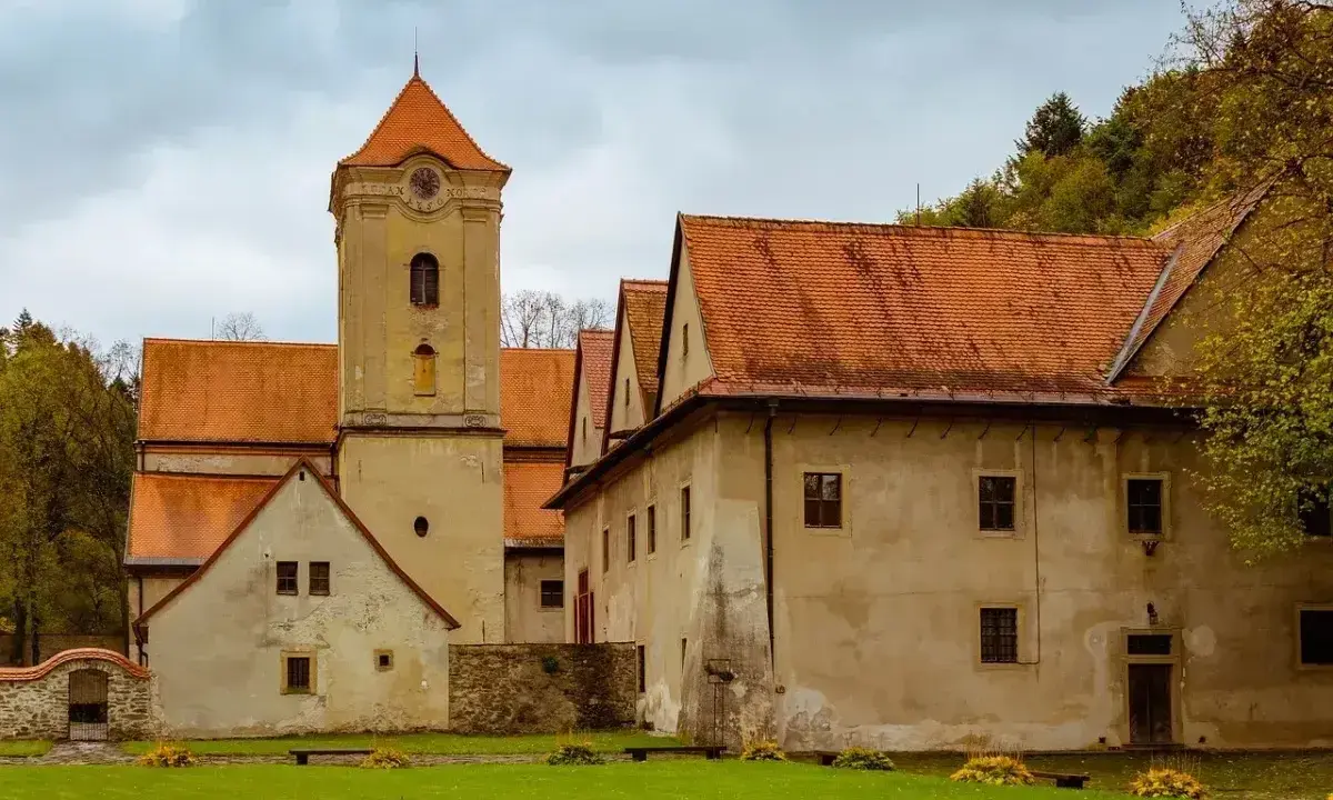 Czerwony klasztor w Pieninach, z charakterystyczną wieżą zegarową i pomarańczowymi dachami, otoczony zielonym trawnikiem i drzewami.