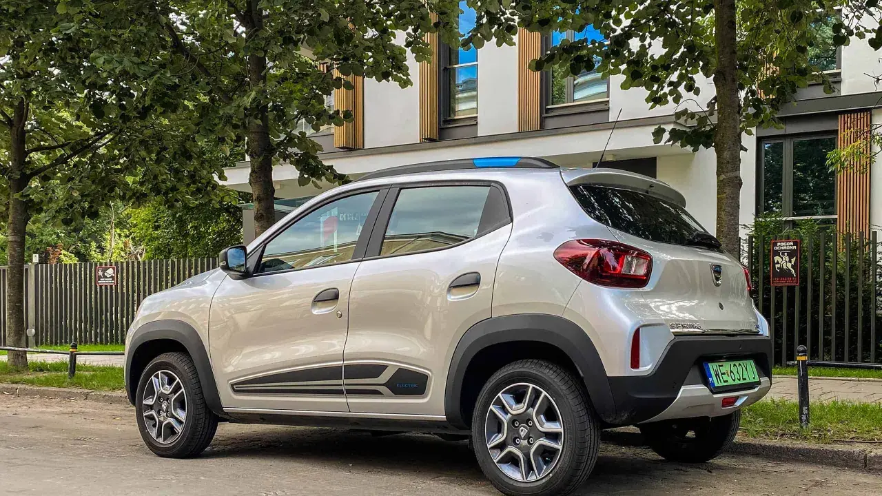 Srebrny Dacia Spring, elektryczny samochód miejski, parkuje pod drzewami. Jego zasięg pozwala na swobodne poruszanie się po mieście.