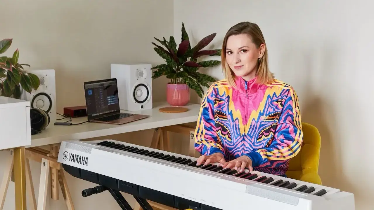 Kobieta gra na pianinie Yamaha. W tle laptop i głośniki, idealne do nauki śpiewu dla dzieci.