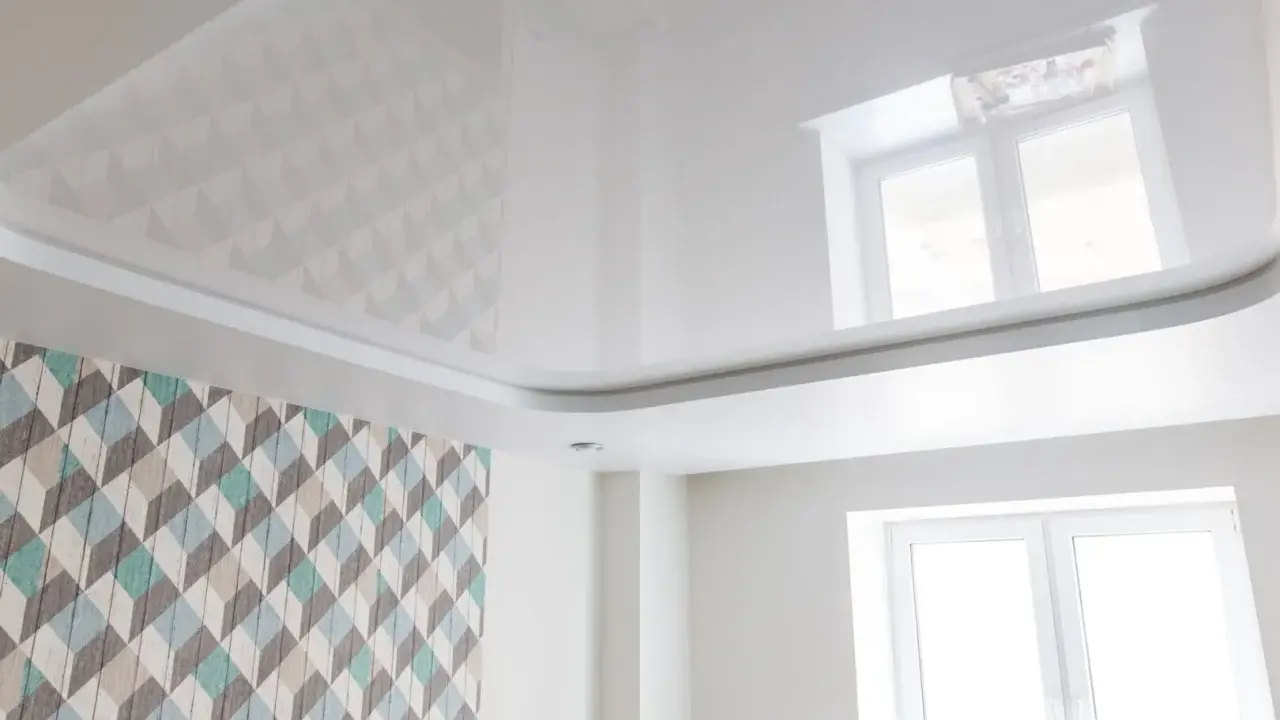 Biały, błyszczący sufit napinany z subtelnymi wzorami geometrycznymi, odbijający światło okna.