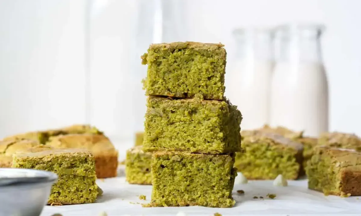 Grüne, saftige matcha brownies, gestapelt und auf einem Tisch arrangiert, mit weißen Schokostückchen und Milchflaschen im Hintergrund.