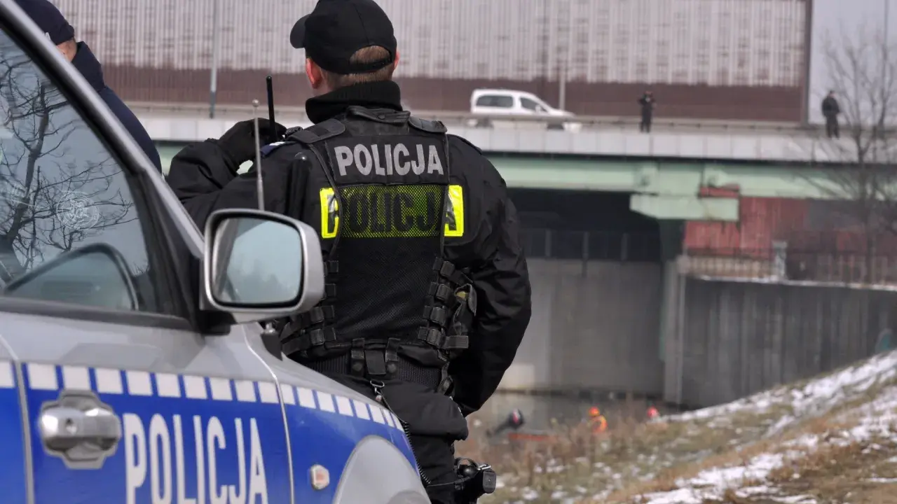 Policjant w kamizelce z napisem "POLICJA", zgodnej z rozporządzeniem w sprawie umundurowania policjantów, stoi obok radiowozu.