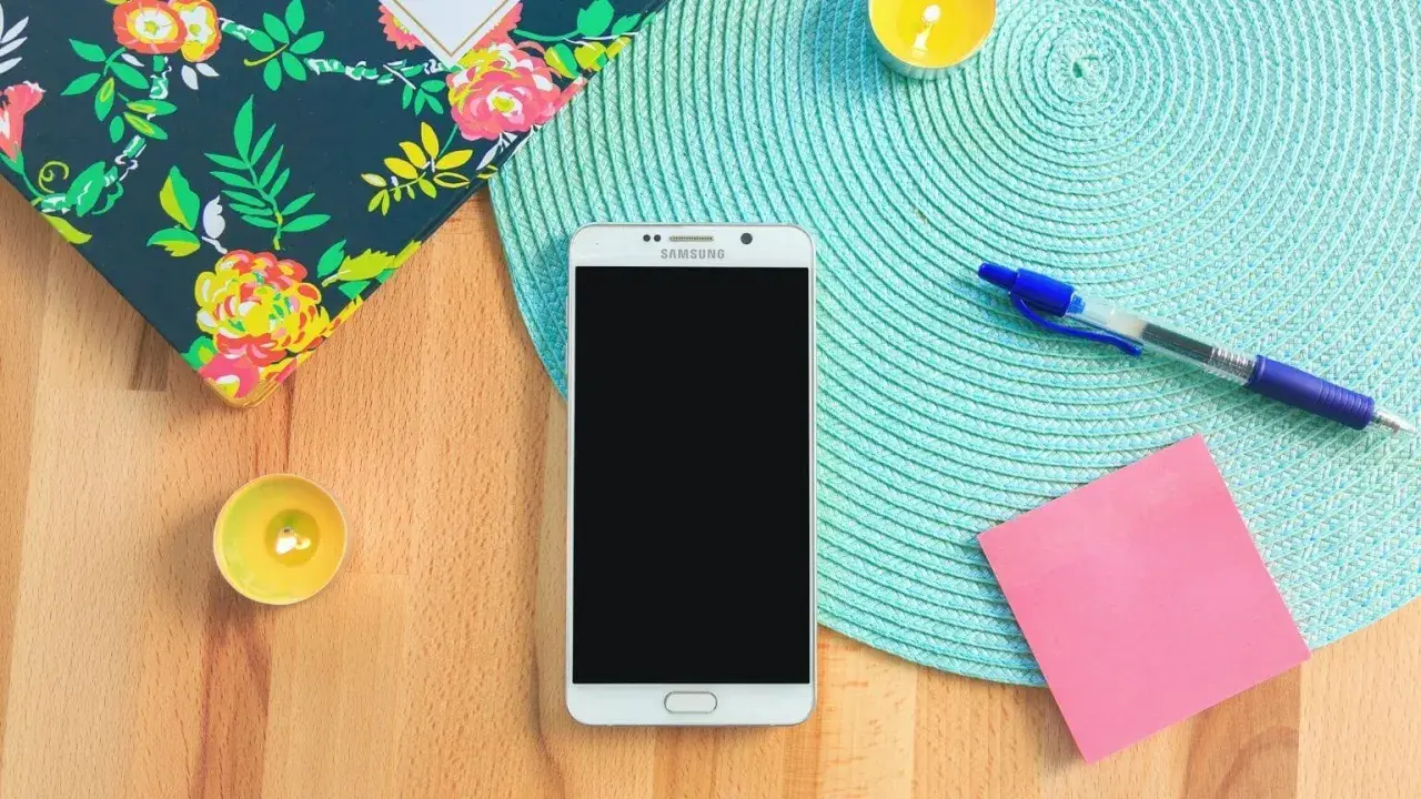 Un smartphone prêt à vous aider à vous organiser au quotidien. Un carnet fleuri, des bougies, un stylo et un post-it rose complètent la scène.