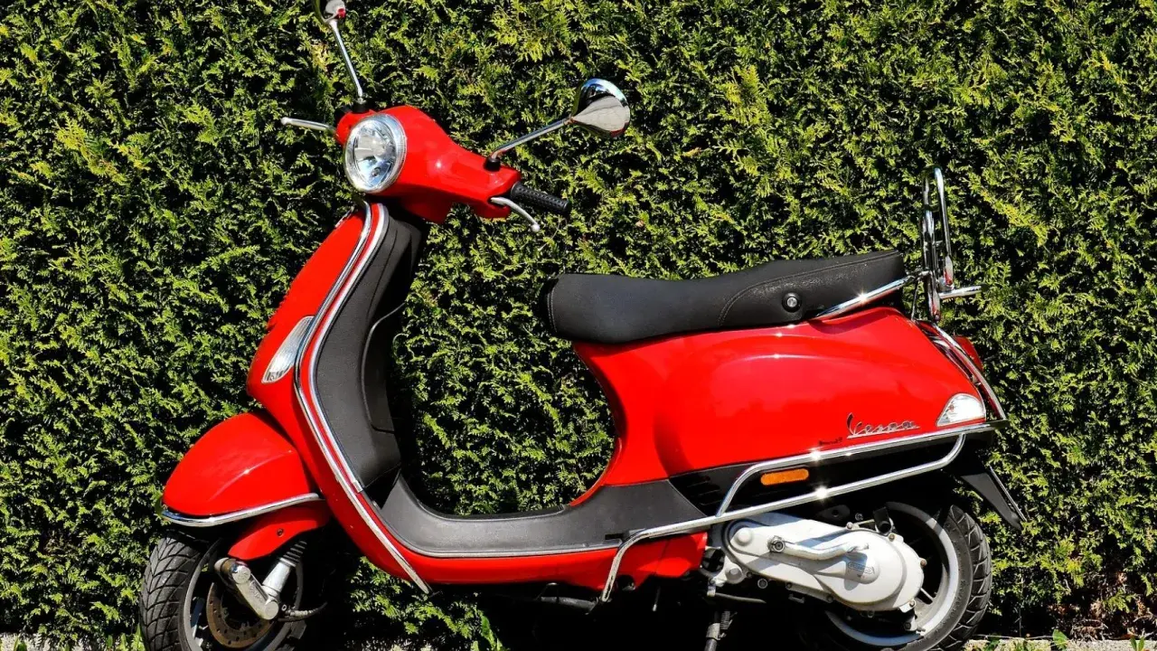 Scooter rojo brillante aparcado frente a un seto verde. ¡Listo para salir con mi permiso AM!