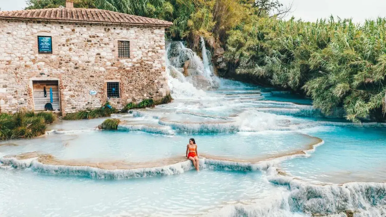 Cascate naturali con acqua turchese, una donna seduta su una roccia. Un antico mulino in pietra sullo sfondo. Tra le terme più belle d'Italia.