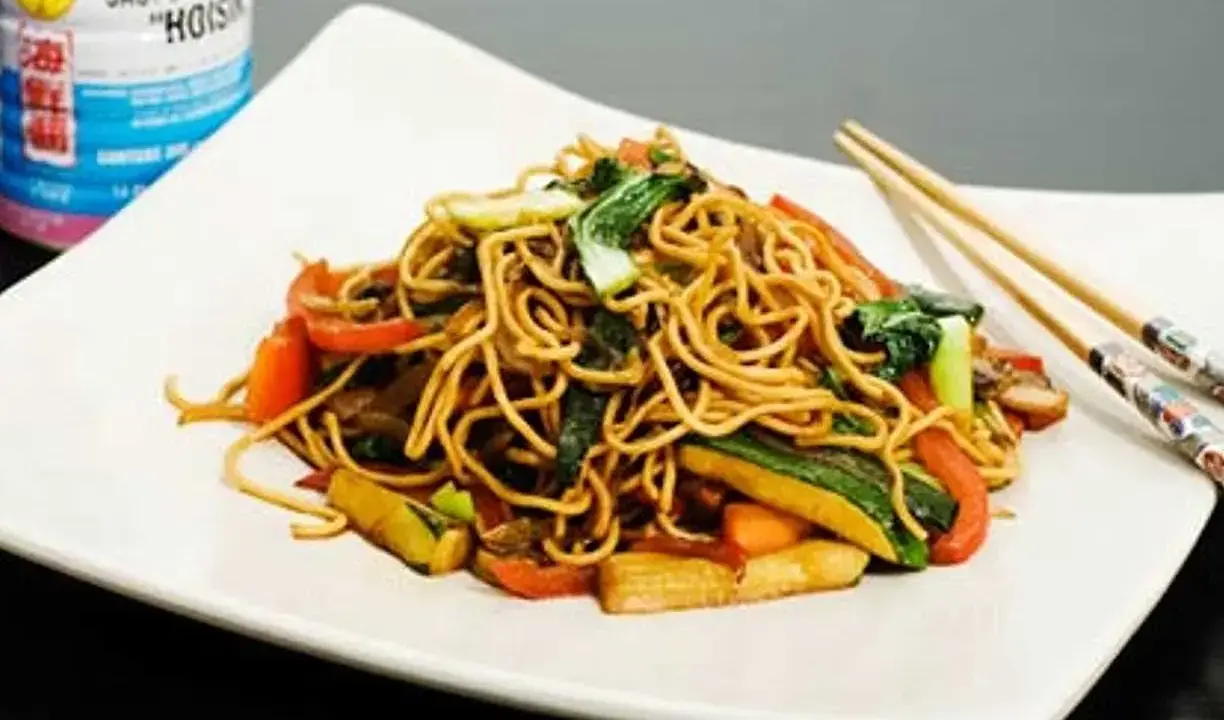En tallrik med nudlar och grönsaker, perfekt för ett snabbt wok recept. Hoisinsåsburk syns i bakgrunden.