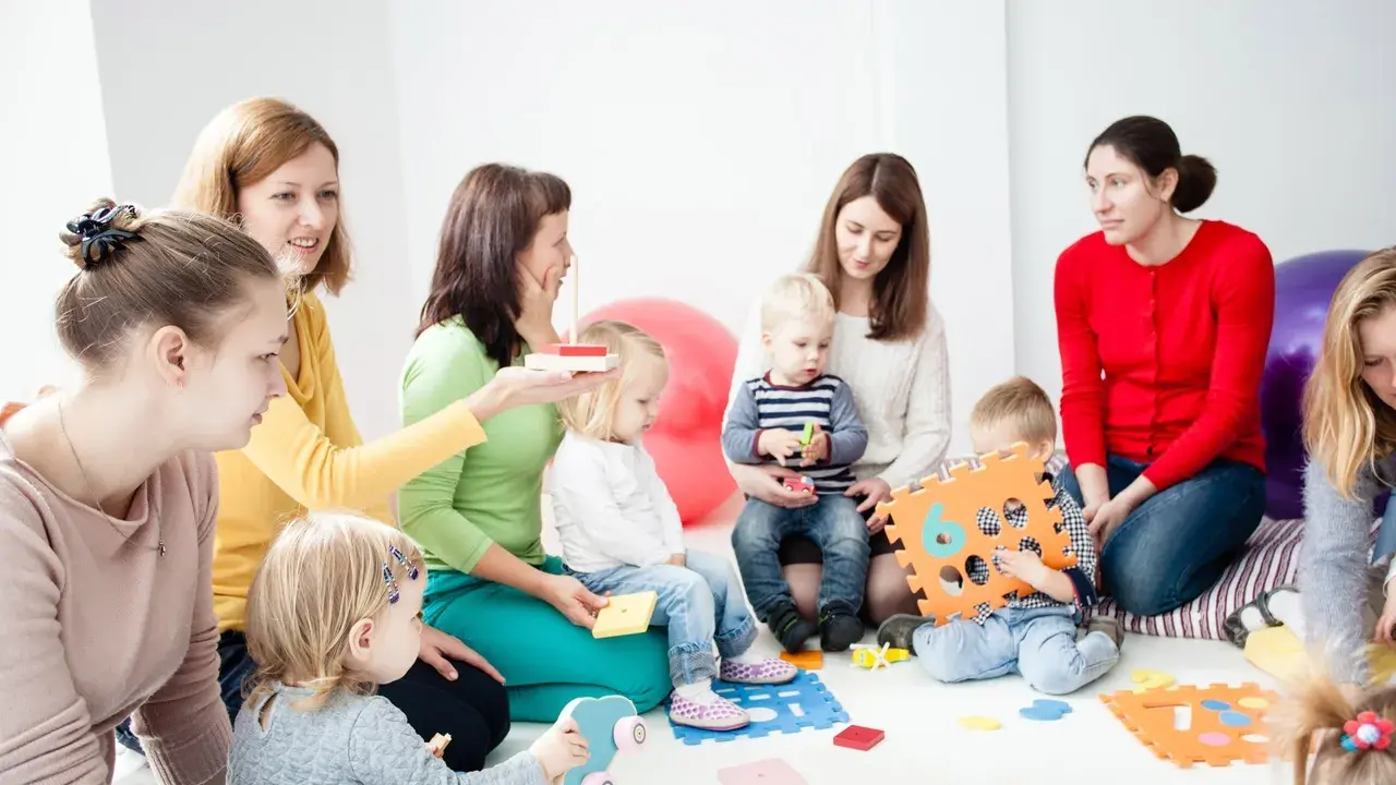 Grupa dzieci i dorosłych uczestniczy w zajęciach, które przypominają terapię pedagogiczną. Dzieci bawią się klockami i zabawkami.