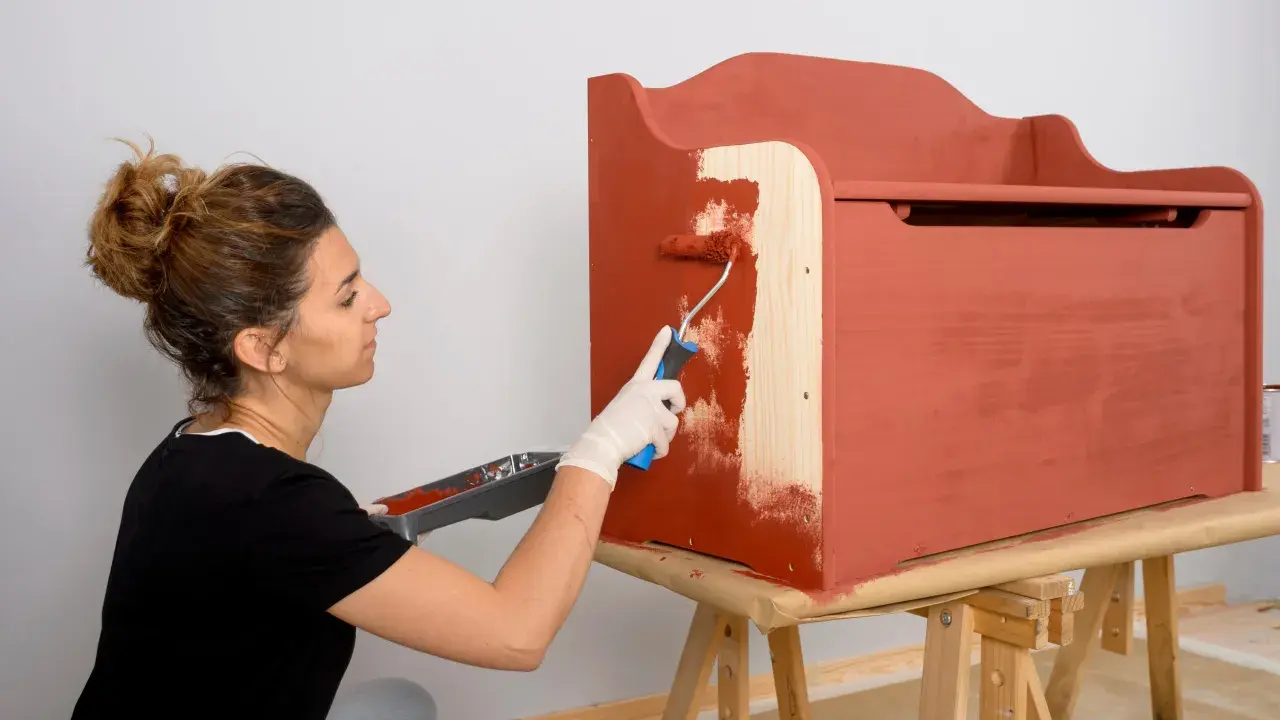 Mujer con guantes blancos pinta con rodillo una caja de madera de color terracota.