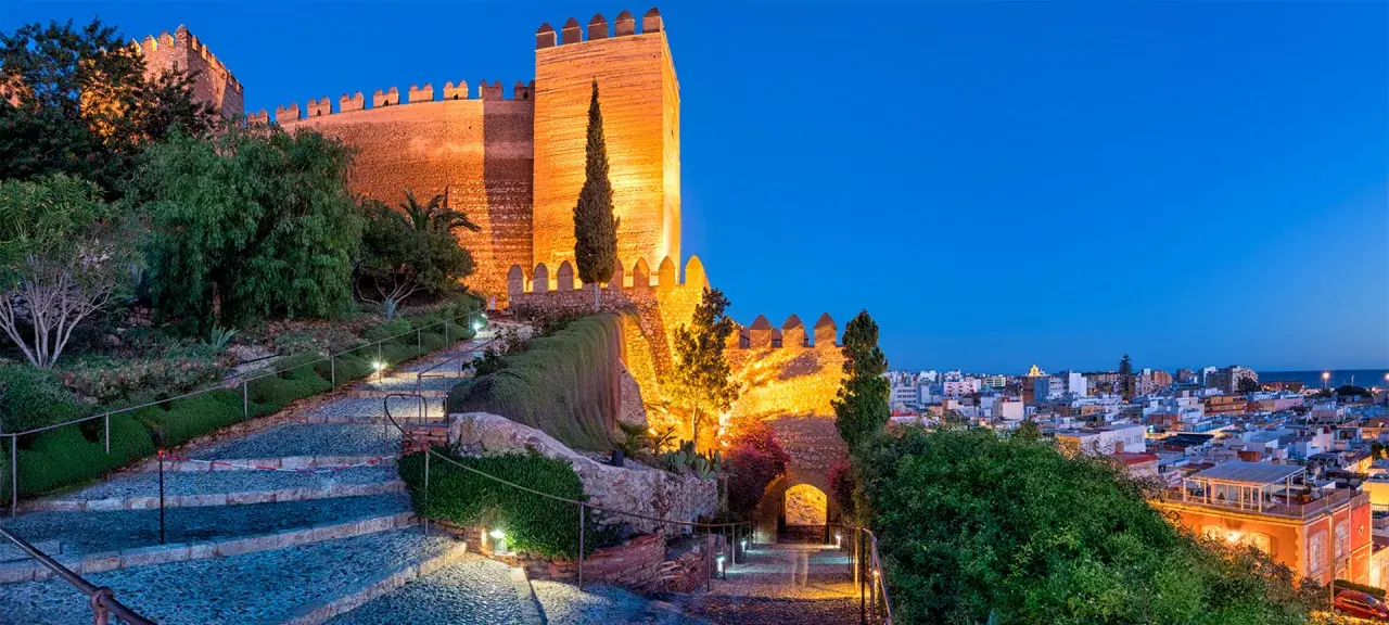 Die Alcazaba von Almería erstrahlt im Abendlicht. Beleuchtete Wege führen durch Gärten zur imposanten Festung.