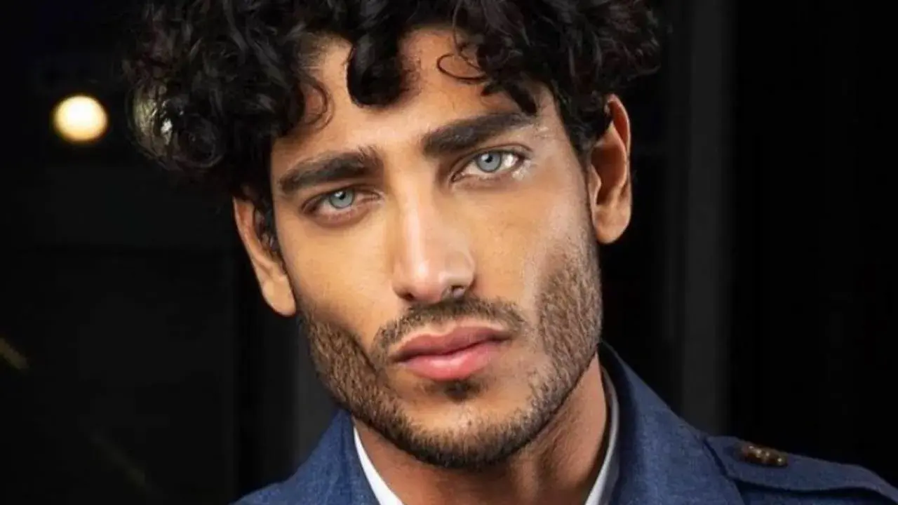 Primo piano di un uomo con splendidi capelli ricci scuri e occhi azzurri penetranti, indossa una giacca blu.