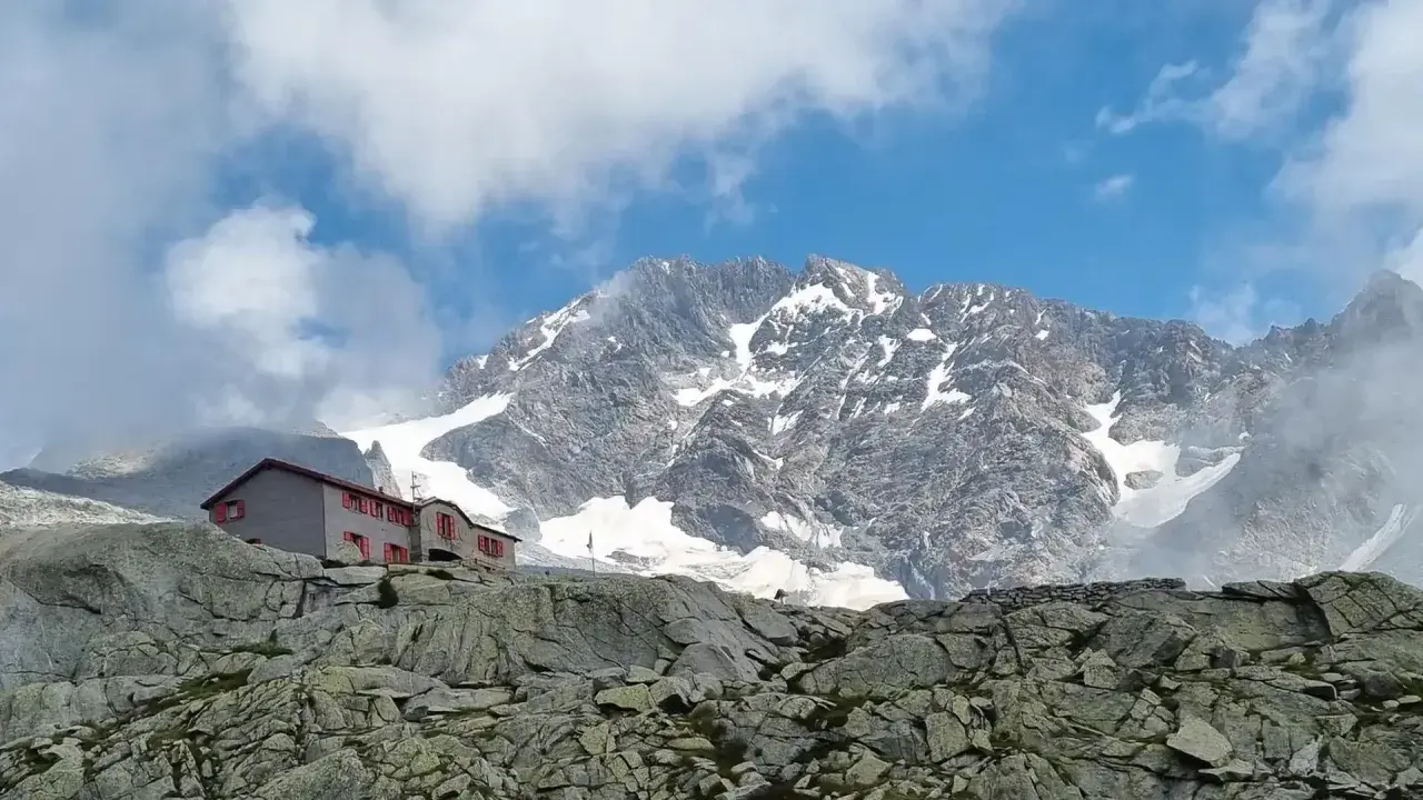 Rifugio Gherardi, un edificio con finestre rosse, si erge su un pendio roccioso sotto un imponente massiccio montuoso innevato.