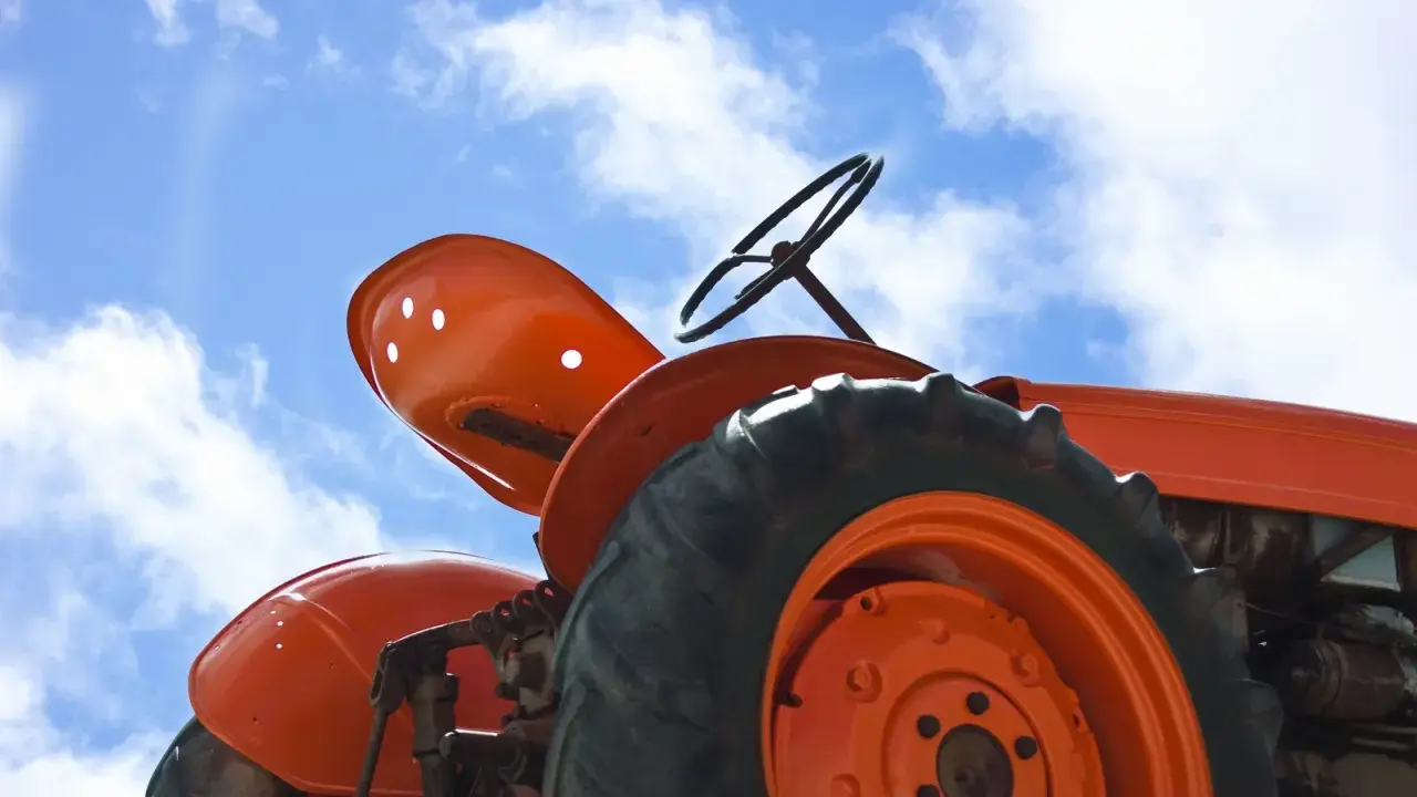 Close-up of an orange tractor's wheel and seat against a blue sky with clouds. This vintage tractor, possibly a different size Kubota tractor, awaits its next task.