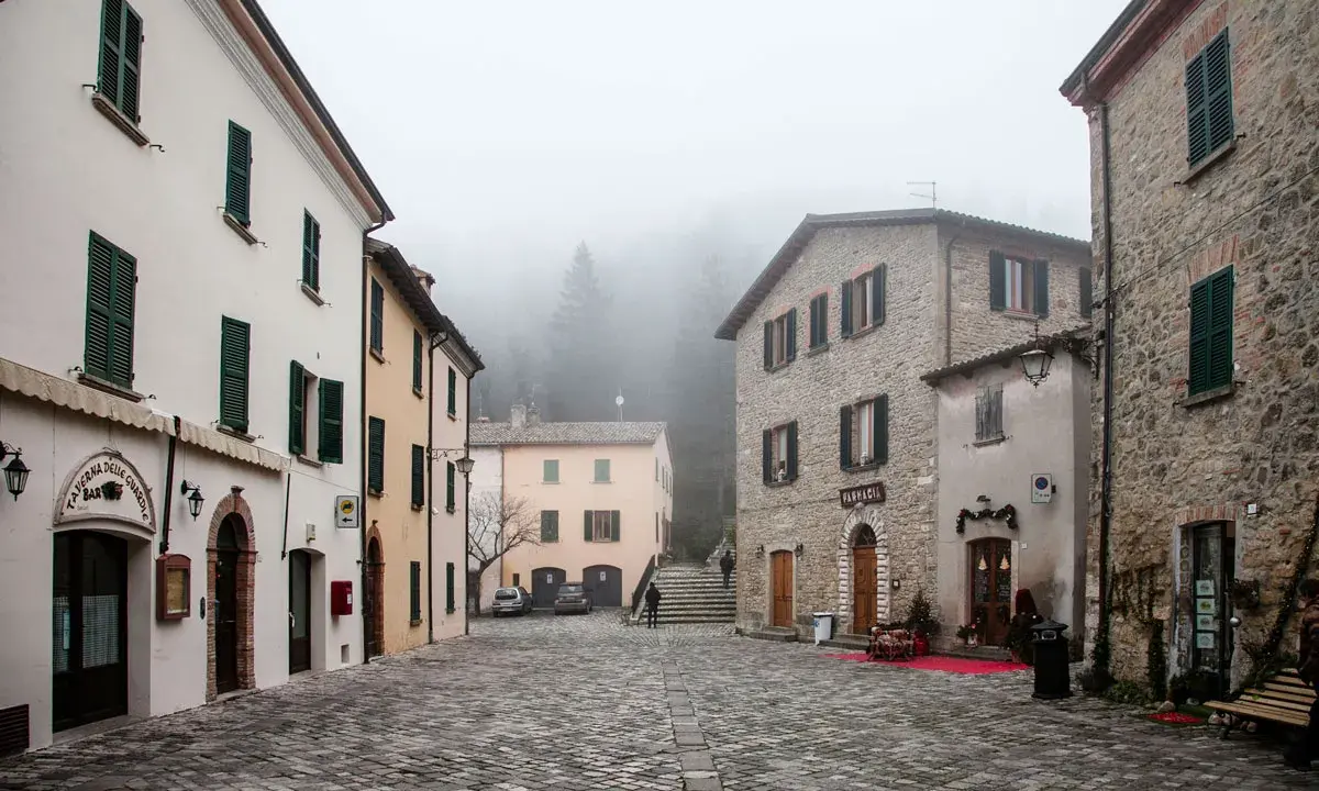 Piazza di San Leo avvolta nella nebbia, con edifici in pietra e intonaco, un bar e una farmacia.