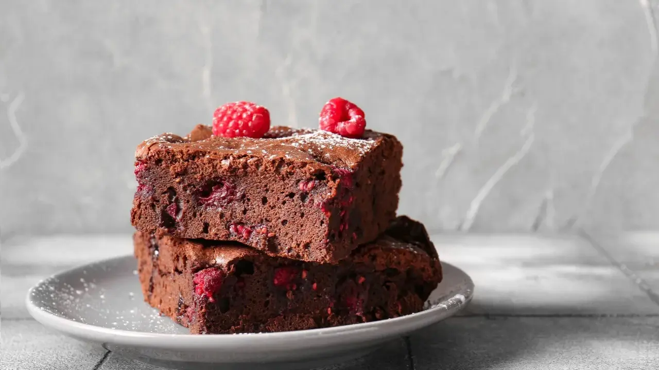 Dwa pyszne brownie z malinami, posypane cukrem pudrem, ułożone na szarym talerzyku.
