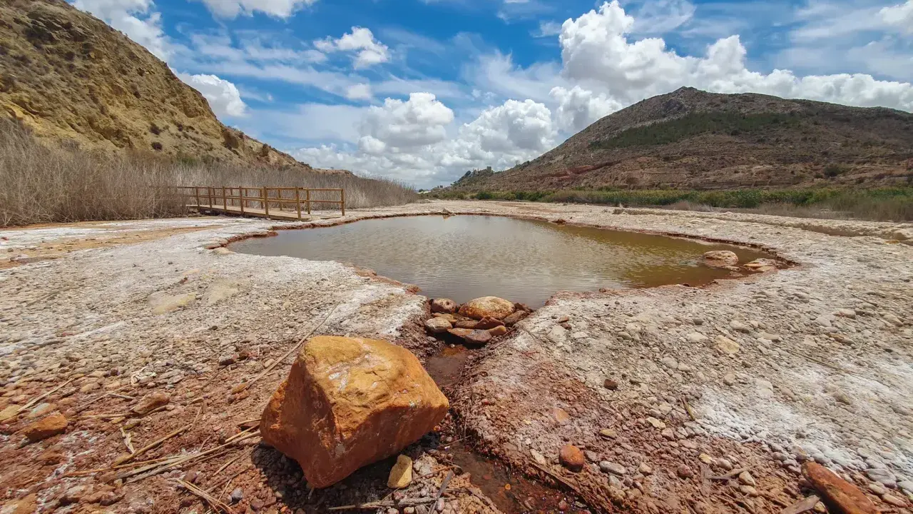 Un estanque natural en Les Salinetes de Novelda, rodeado de rocas y vegetación seca, bajo un cielo azul con nubes blancas.