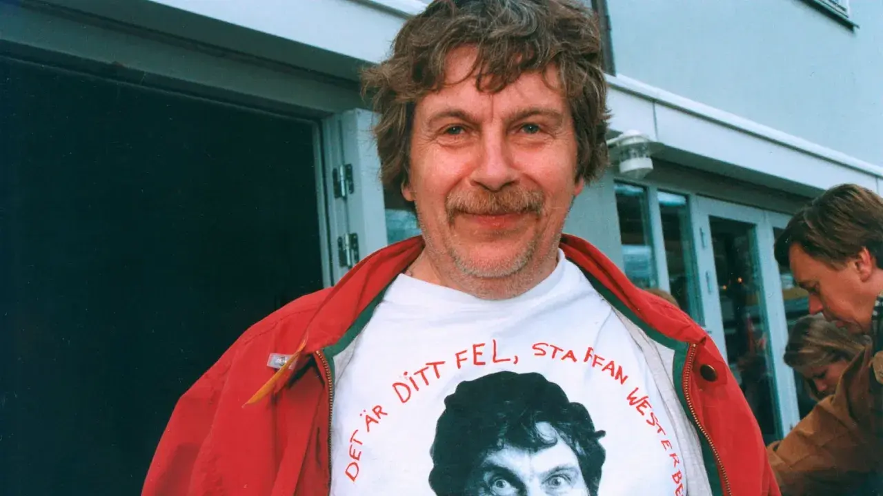 Staffan Westerberg, känd för sina filmer och tv-program, ler i en röd jacka över en t-shirt med hans eget ansikte och texten "Det är ditt fel, Staffan Westerberg".