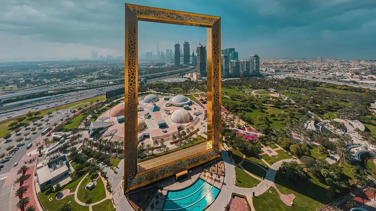 Dubai Frame rahmt die Skyline und die futuristischen Dubai Projekte ein. Ein Park mit Kuppeln und ein Pool sind im Vordergrund.