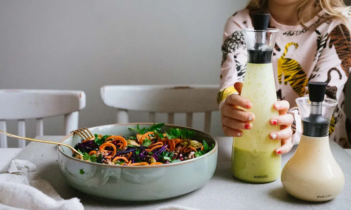 En skål med färgglad sallad och två flaskor dressing till sallad. En flicka håller i den ena flaskan.