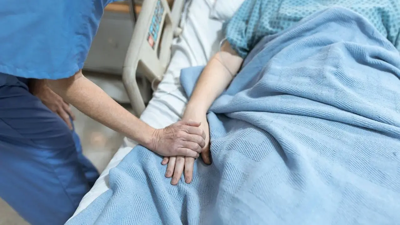 Pielęgniarka trzyma za rękę pacjentkę leżącą w łóżku szpitalnym pod niebieskim kocem.