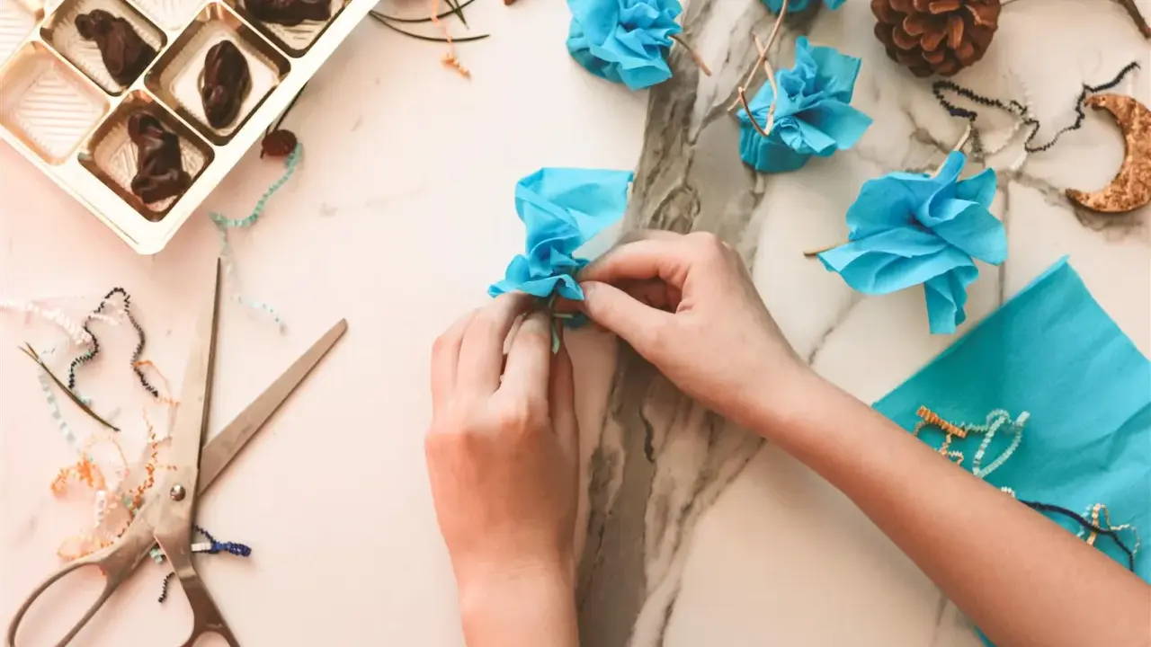 Ręce tworzą niebieskie kwiatki z papieru, pokazując, co można zrobić z cienkiej wstążki. Obok czekoladki i nożyczki.