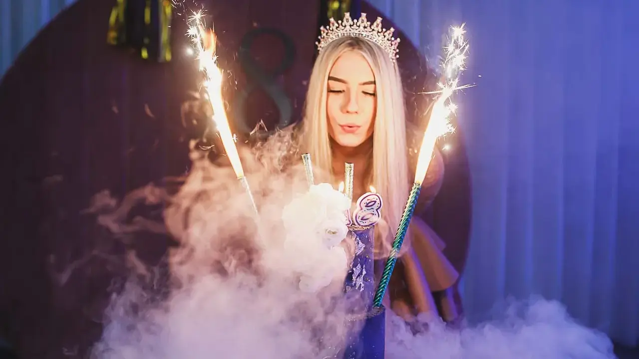 Dziewczyna w koronie zdmuchuje świeczki na torcie z fajerwerkami.
