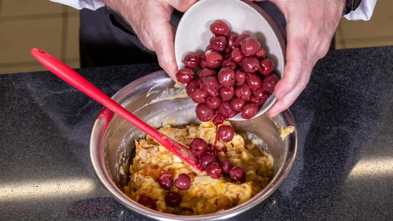 Hände geben Kirschen in eine Schüssel mit Teig für ein einfaches Kirschenmichel Rezept. Ein roter Teigschaber liegt daneben.