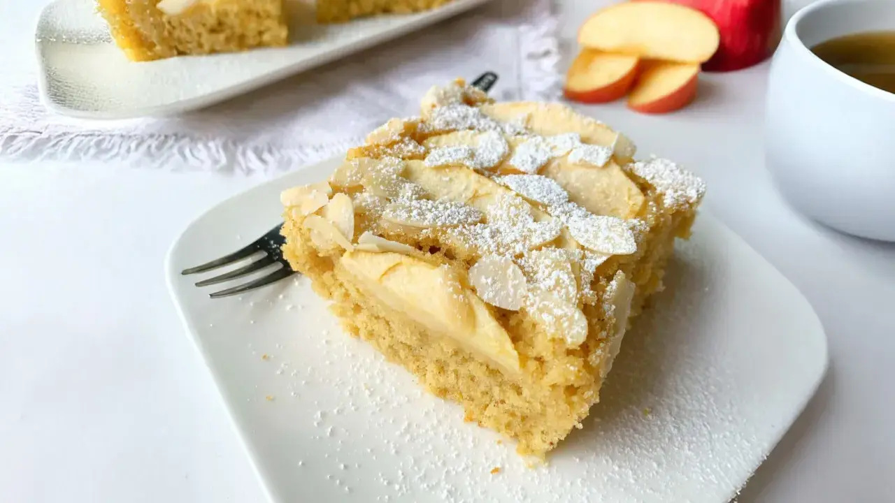 Ein Stück saftiger Apfelkuchen mit Apfelmus, bestreut mit Puderzucker und Mandelblättchen, wartet auf dem Teller.