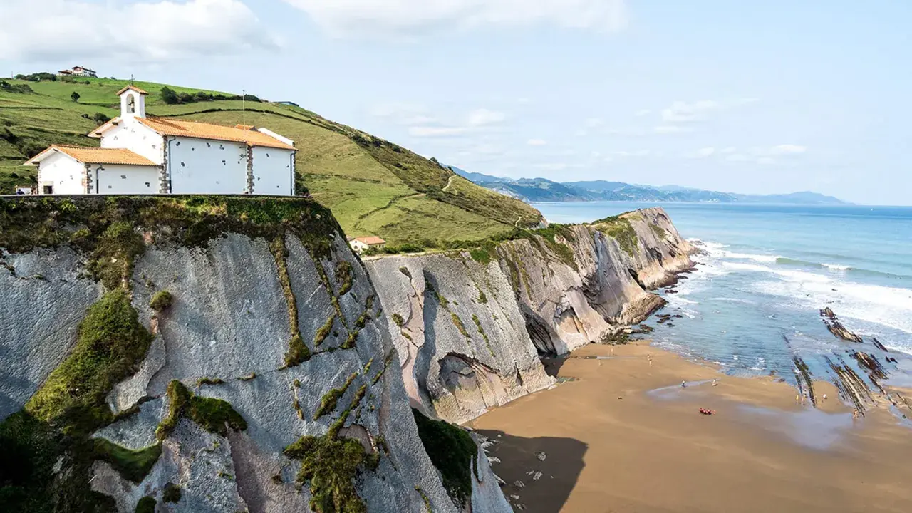 Ermita blanca sobre acantilados en la playa de Itzurun, con el mar Cantábrico y olas rompiendo.