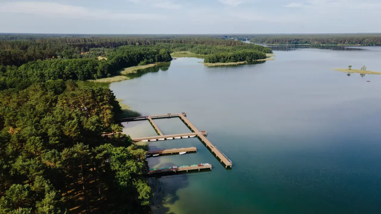 Drewniane pomosty na spokojnym jeziorze Wiartel, otoczonym gęstym lasem. Woda jest przejrzysta, a na pomostach stoją łódki i kajaki.