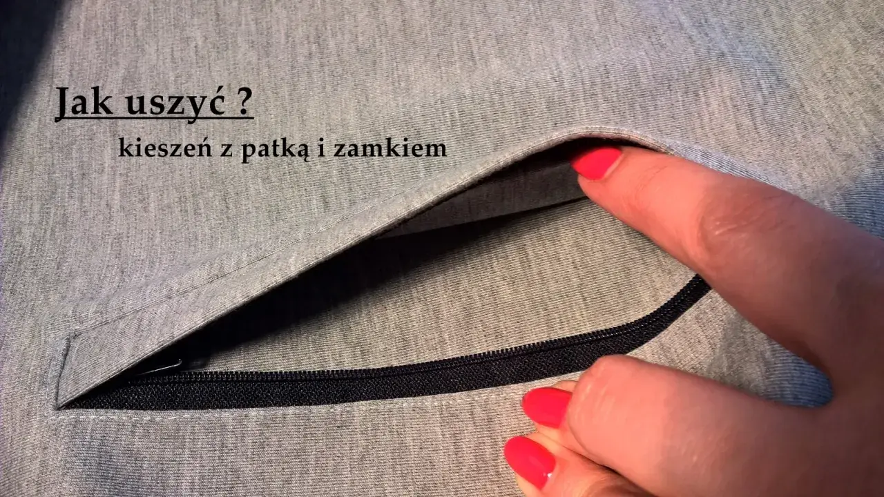Szara tkanina z widocznym zamkiem i patką. Dłoń z czerwonym lakierem do paznokci wsuwa się do kieszeni z patką.