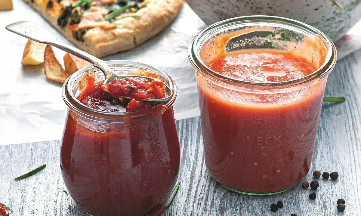 Dwa słoiczki domowego sosu do pizzy, jeden otwarty z łyżeczką, obok kawałek pizzy i przyprawy.