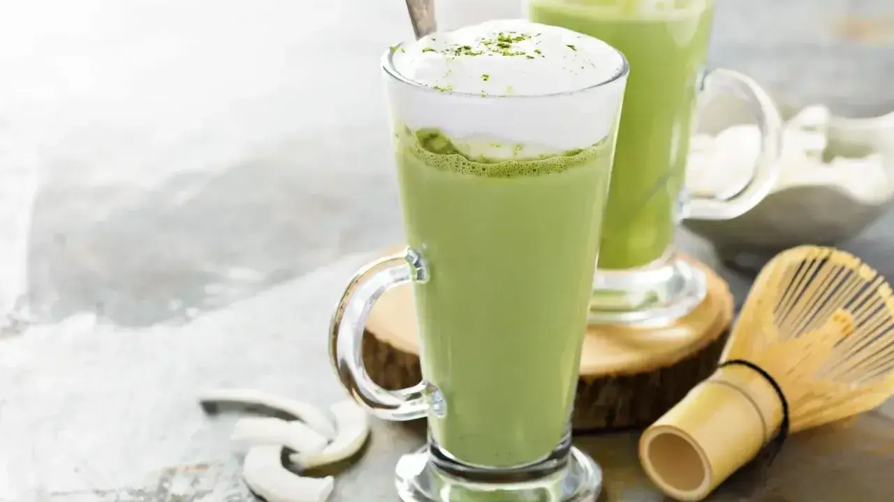 Dwa zielone napoje matcha w szklankach z pianką, obok bambusowa miotełka i wiórki kokosowe. Tak smakuje matcha.