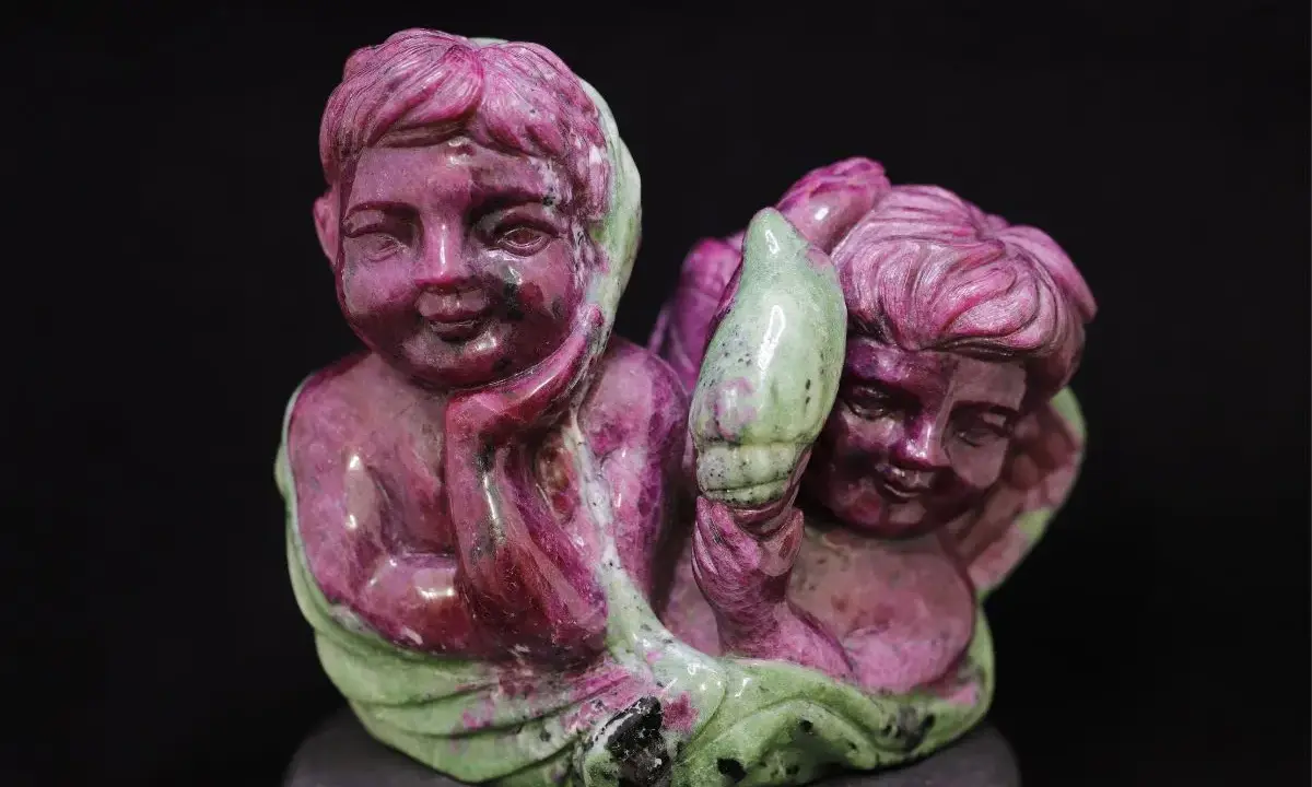 Due putti scolpiti in pietra, uno con la mano sul mento, sembrano abbracciare un elemento verde, forse un fiore. La pietra, un rubino verde, dona un tocco di magia.
