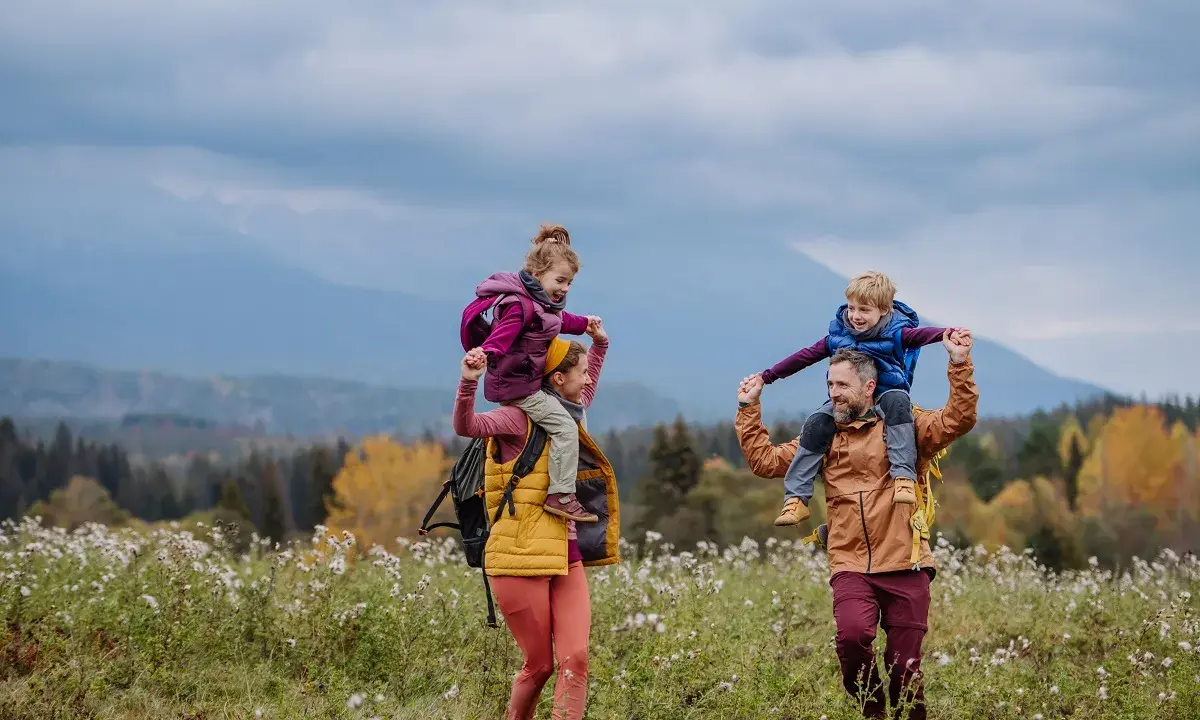 Rodzina na spacerze po łące. Dzieci siedzą na ramionach rodziców, jakby hasające zające zakopane w trawie.