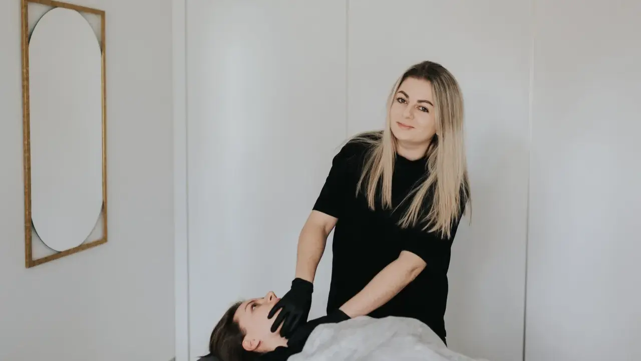 Terapeuta w czarnych rękawiczkach wykonuje zabieg rehabilitacji głosu u leżącej pacjentki.