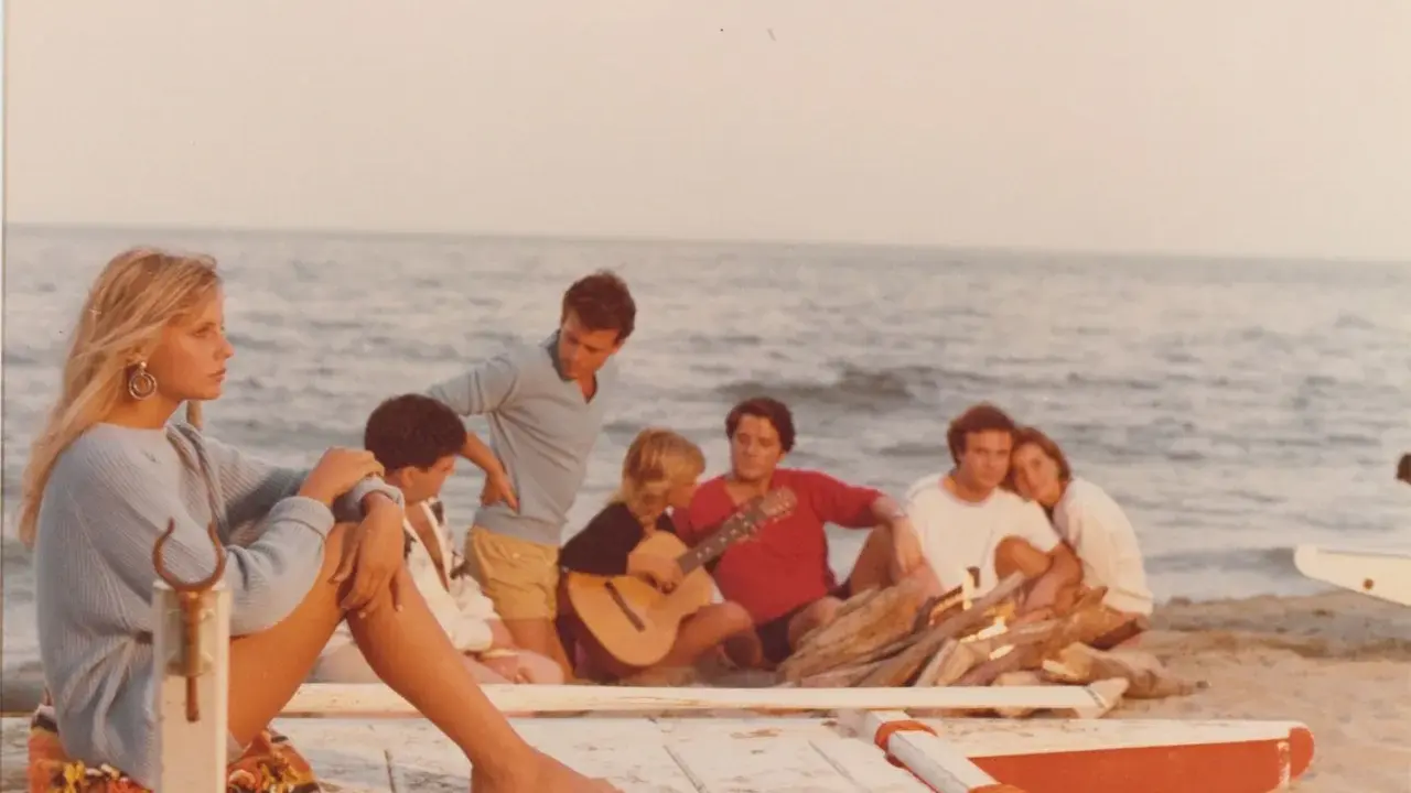 Giovani su una spiaggia, un gruppo di amici che ricorda i film estivi italiani, con chitarra e falò.