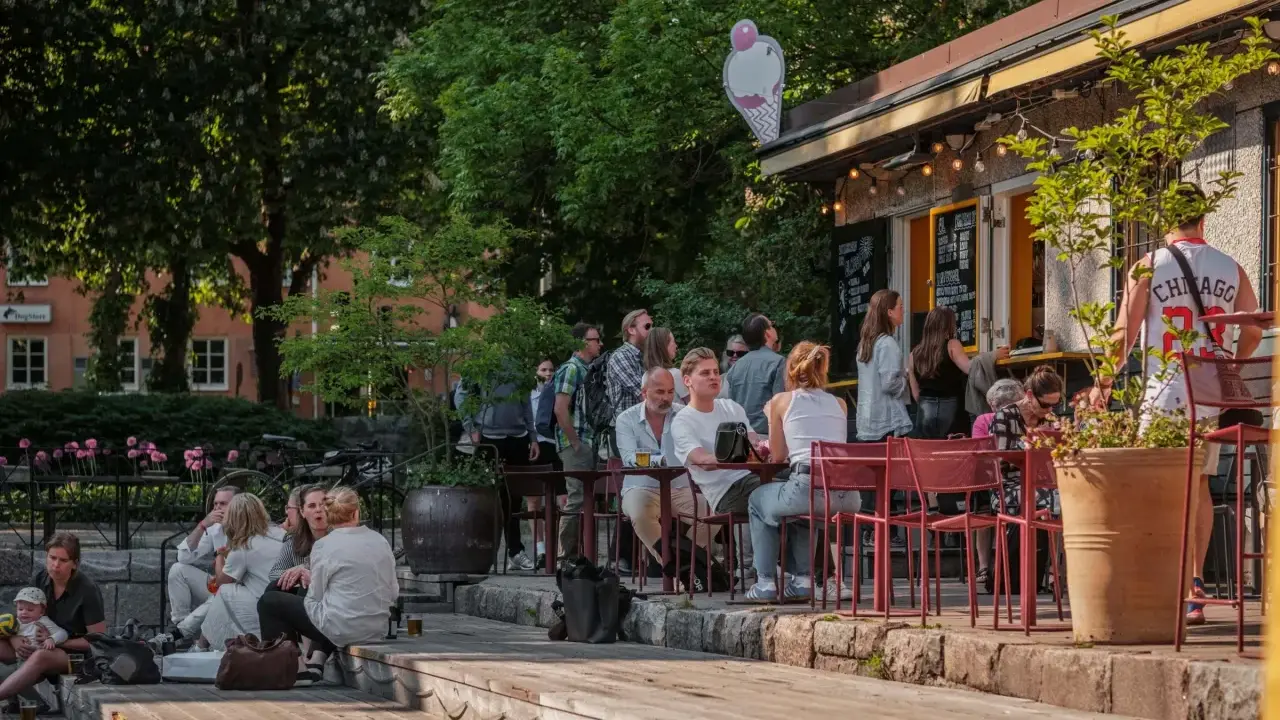 Folk njuter av solen på uteserveringar i Stockholm. En glasskylt syns ovanför en servering.