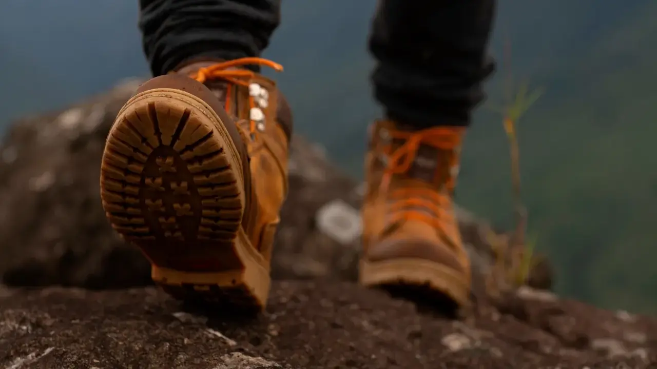 Stopy w brązowych butach trekkingowych z pomarańczowymi sznurówkami na kamienistym szlaku. Membrana w butach zapewnia komfort podczas wędrówki.
